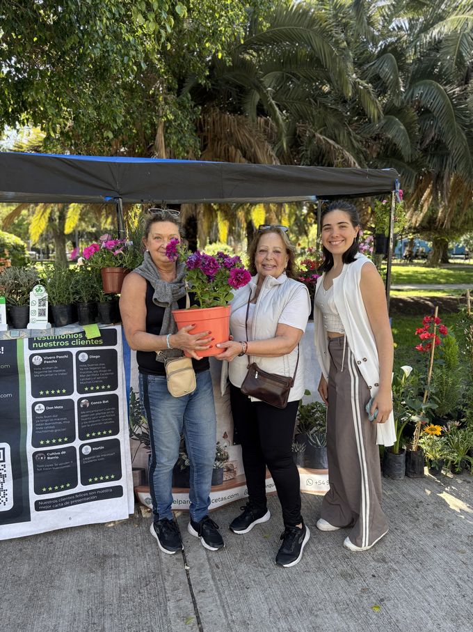 Tres clientas sonrientes frente al puesto de El Parque Macetas; una sostiene una maceta con flores
