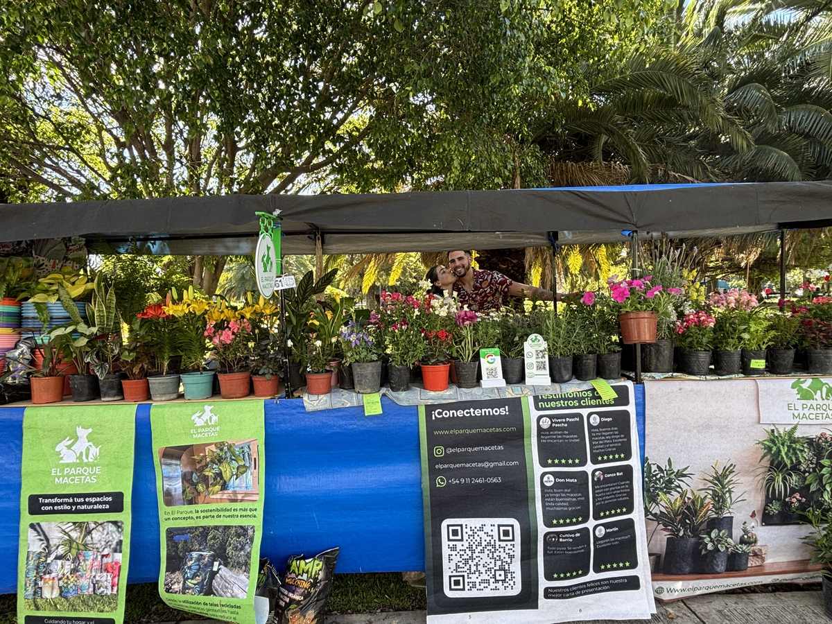 El puesto de El Parque Macetas en la feria: plantas con flores en fila sobre el mostrador, carteles de la marca y dos personas detrás