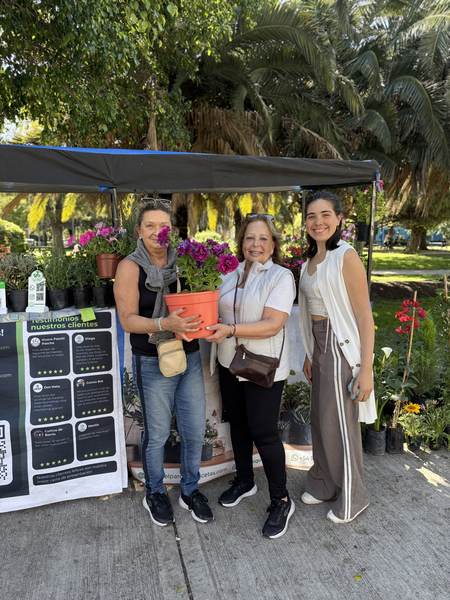 Tres mujeres sonrientes con una maceta con flores en el puesto de El Parque Macetas en la feria